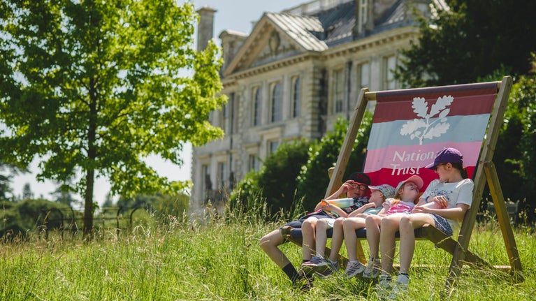 Four children in summer clothes sitting in a giant deckchair in front of a stone mansion. It is a sunny day and the grass is long